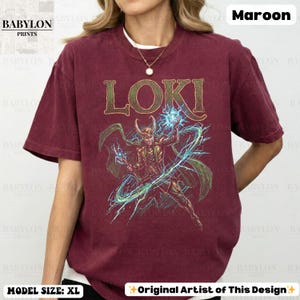 May include: Maroon t-shirt featuring the word "LOKI" in gold lettering above a detailed illustration of a character with a horned helmet, holding a glowing blue orb. The shirt is from Babylon Prints and the model is wearing size XL.