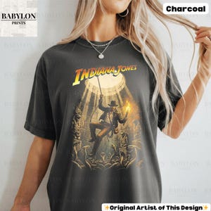 May include: Charcoal-colored t-shirt featuring the Indiana Jones movie logo and a graphic of the character in an action scene. The shirt has a vintage look, with the text "Babylon Prints" in the upper left corner and "Original Artist of This Design" at the bottom.