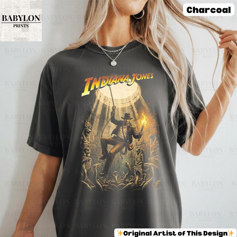 May include: Charcoal-colored t-shirt featuring the Indiana Jones movie logo and a graphic of the character in an action scene. The shirt has a vintage look, with the text "Babylon Prints" in the upper left corner and "Original Artist of This Design" at the bottom.