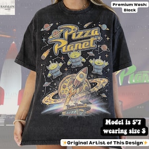 May include: Black t-shirt featuring a colorful graphic design with the text "Pizza Planet". The design includes aliens, a rocket ship, planets, and a pizza. The shirt is a premium wash.