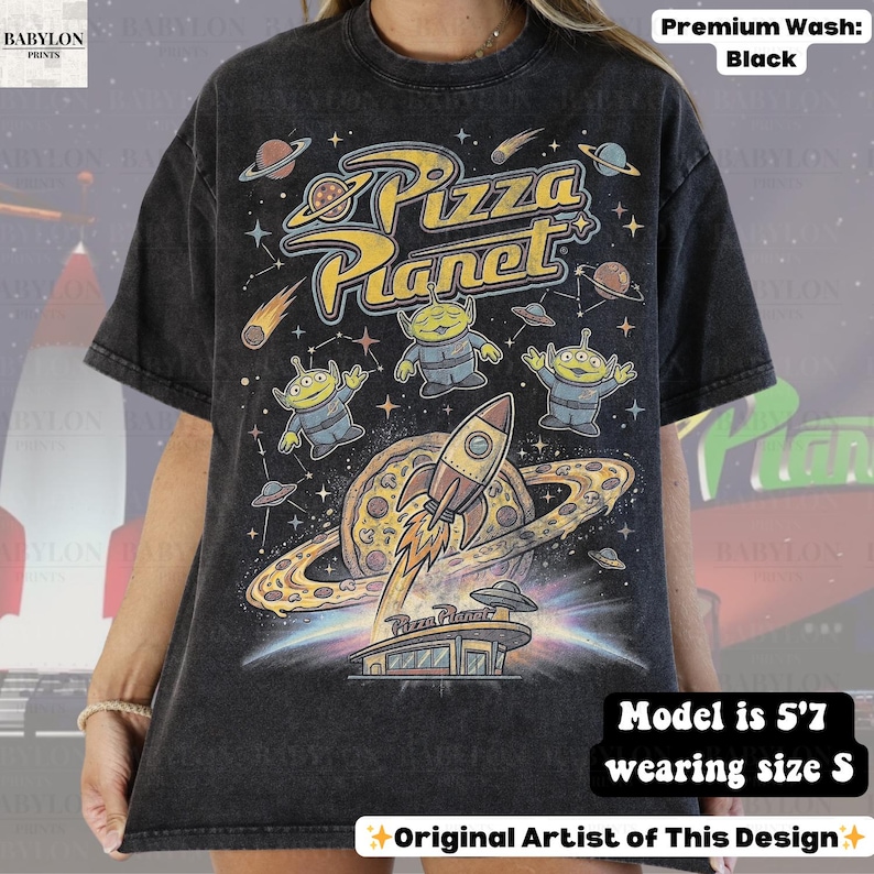 May include: Black t-shirt featuring a colorful graphic design with the text "Pizza Planet". The design includes aliens, a rocket ship, planets, and a pizza. The shirt is a premium wash.