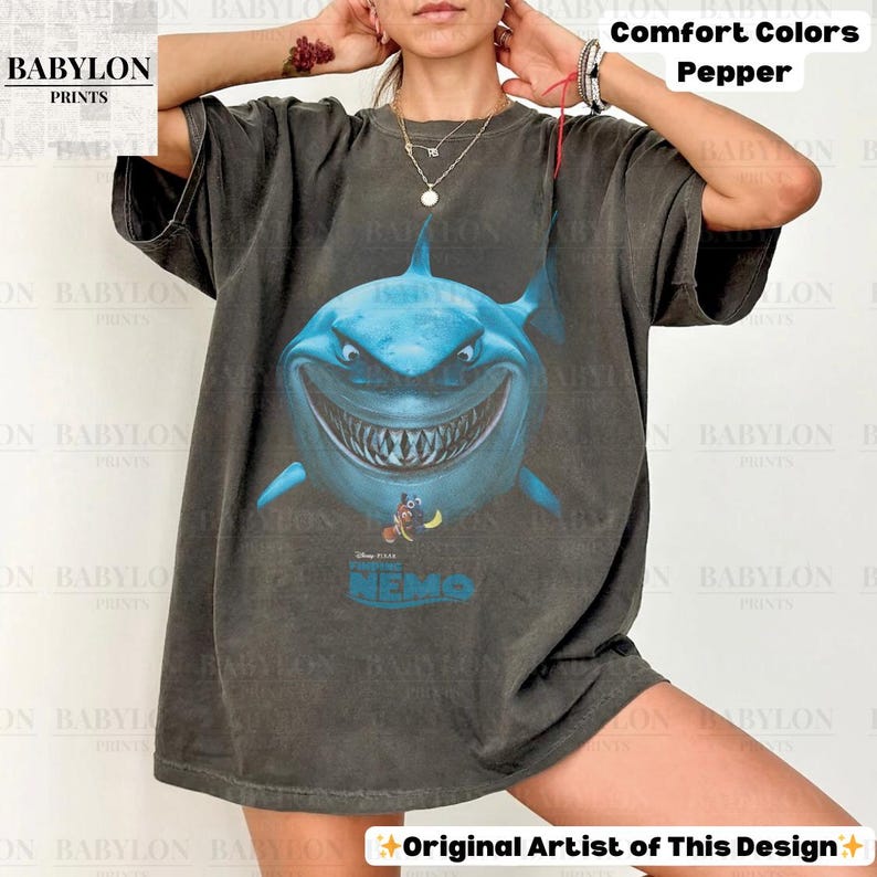 May include: A dark gray t-shirt featuring a large, blue cartoon shark with a menacing grin. The shirt has the text "NEMO" and the Disney Pixar logo. The words "Comfort Colors Pepper" are printed on the top right. The Babylon Prints logo is on the top left.
