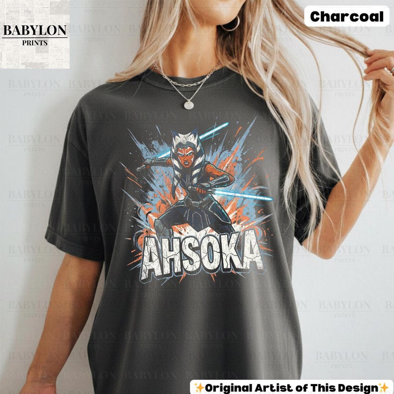 Może przedstawiać: Szary t-shirt z nadrukiem Ahsoki Tano z mieczami świetlnymi i napisem "AHSOKA". Koszulka ma kr&oacute;tkie rękawy i okrągły dekolt. Widoczne jest logo Babylon Prints. Widoczny jest r&oacute;wnież napis "Charcoal".