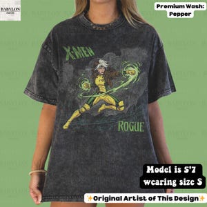 May include: A dark grey t-shirt with a graphic of the X-Men character Rogue in a yellow and green costume. The text "X-MEN" and "ROGUE" are printed on the shirt. The shirt is a premium wash, pepper colour.