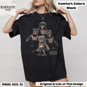 Op de afbeelding: Een zwart t-shirt met korte mouwen en een grafische print van vier Ewok-personages uit Star Wars. Het ontwerp toont de wezens in bruine en beige tinten met het Star Wars-logo in witte tekst. Het shirt is gelabeld als Comfort Colors Black.