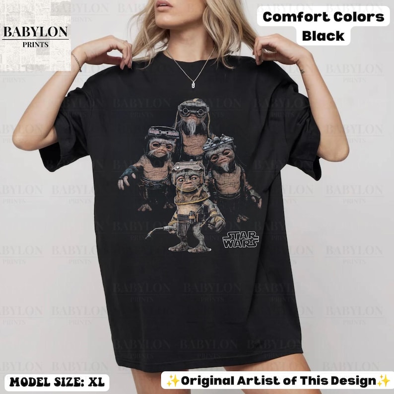 Op de afbeelding: Een zwart t-shirt met korte mouwen en een grafische print van vier Ewok-personages uit Star Wars. Het ontwerp toont de wezens in bruine en beige tinten met het Star Wars-logo in witte tekst. Het shirt is gelabeld als Comfort Colors Black.