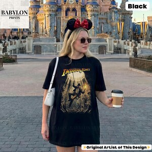 May include: Black t-shirt featuring the text "INDIANA JONES" and a graphic of the character. The shirt is from "BABYLON PRINTS" and has "Original Artist of This Design" text. The shirt is worn by a person in front of a castle.