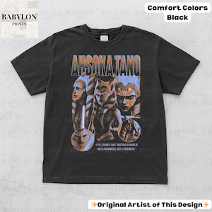 May include: Black t-shirt with a vintage-style graphic of Ahsoka Tano from Star Wars. The graphic features Ahsoka and other characters from the show. The text "I've learned that trusting others is not a weakness, but a strength." is printed below the graphic.