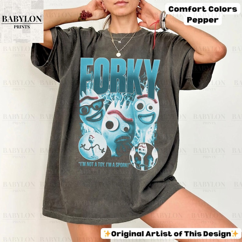 May include: Dark grey t-shirt featuring the word "FORKY" in large, teal letters above a graphic of three Forky characters. The shirt includes the text "I'M NOT A TOY. I'M A SPORK!" and is labeled "Comfort Colors Pepper."