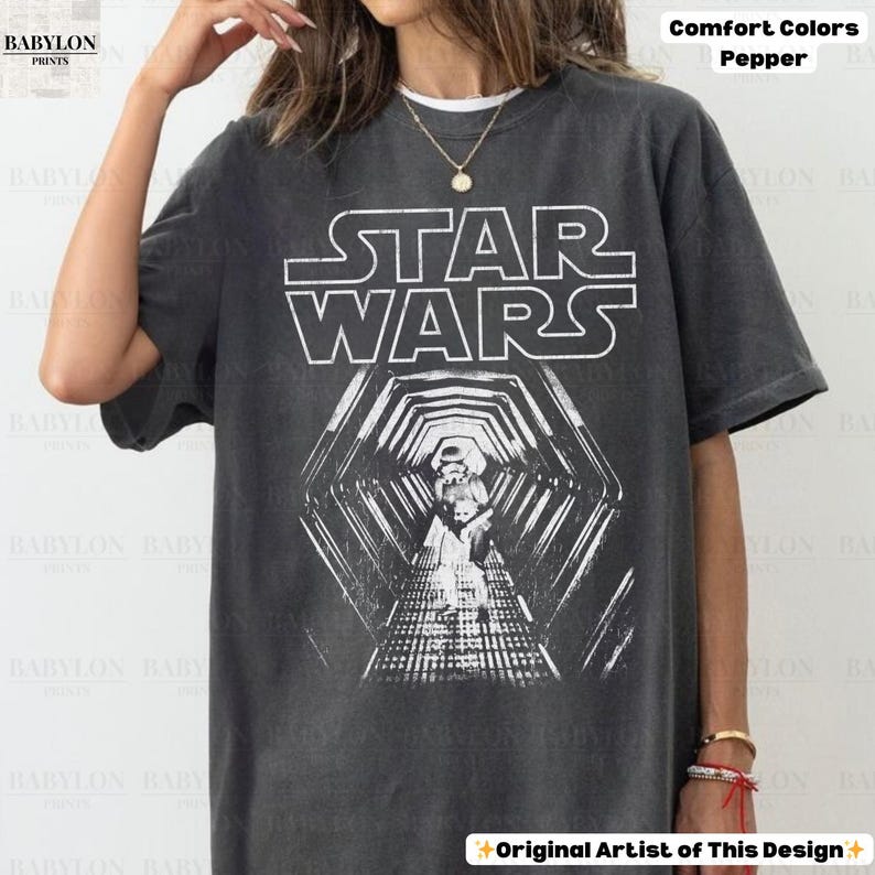 May include: Dark grey t-shirt featuring the Star Wars logo in white, with a graphic of a tunnel and Stormtroopers. The shirt is labeled "Comfort Colors Pepper" and "Original Artist of This Design."