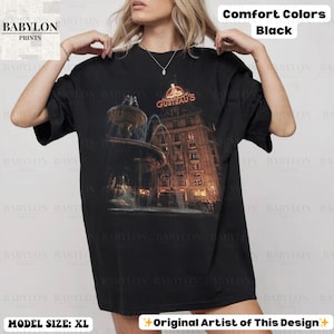 May include: Black comfort colours t-shirt featuring a building and fountain graphic, with the text "GUSTEAU'S". The shirt is size XL. Design by the original artist.