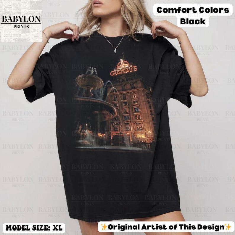 May include: Black comfort colours t-shirt featuring a building and fountain graphic, with the text "GUSTEAU'S". The shirt is size XL. Design by the original artist.