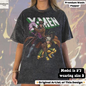 May include: A pepper-wash, charcoal-colored t-shirt featuring the X-Men logo in green and white. The graphic design includes illustrations of Gambit and Rogue. The model is 5'7" and wearing a size S. The design is by the original artist.