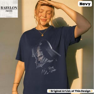 K&ouml;nnte beinhalten: Marineblaues T-Shirt mit einer schwarz-wei&szlig;en Illustration von Darth Vader. Der Aufdruck enth&auml;lt den Text "Darth Vader". Das T-Shirt hat einen Rundhalsausschnitt und kurze &Auml;rmel. Das Babylon Prints Logo ist in der oberen linken Ecke sichtbar.