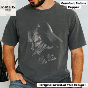 K&ouml;nnte beinhalten: Dunkelgraues T-Shirt mit einer schwarz-wei&szlig;en Illustration von Darth Vader. Der Text "Darth Vader" ist kursiv geschrieben. Das Shirt tr&auml;gt auch die Aufschrift "Comfort Colors Pepper" und "Original Artist of This Design".