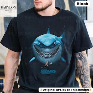 May include: Black t-shirt featuring a large, detailed illustration of a blue shark with a wide grin. Below the shark is the text "Finding Nemo" and the Disney Pixar logo. The shirt also has the text "Original Artist of This Design".