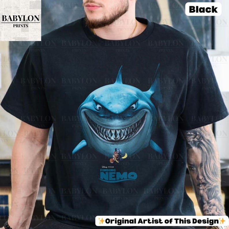 May include: Black t-shirt featuring a large, detailed illustration of a blue shark with a wide grin. Below the shark is the text "Finding Nemo" and the Disney Pixar logo. The shirt also has the text "Original Artist of This Design".