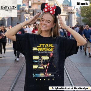 Puede incluir: Camiseta negra de Star Wars con el texto "STAR WARS THE PHANTOM MENACE" en amarillo y un gr&aacute;fico de una batalla de sables de luz. La camiseta es de estilo cuello redondo cl&aacute;sico.