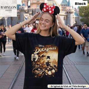May include: Black t-shirt featuring a vintage-style "DuckTales" graphic. The design includes characters like Scrooge McDuck and Huey, Dewey, and Louie. The shirt has a classic fit and is made of a soft material. The text "Original Artist of This Design" is visible.