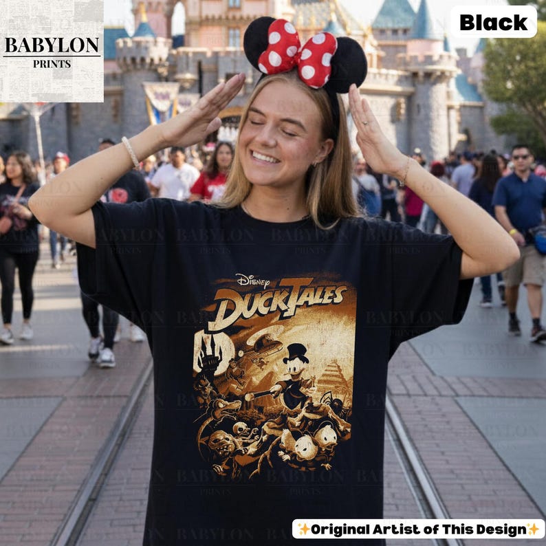 May include: Black t-shirt featuring a vintage-style "DuckTales" graphic. The design includes characters like Scrooge McDuck and Huey, Dewey, and Louie. The shirt has a classic fit and is made of a soft material. The text "Original Artist of This Design" is visible.