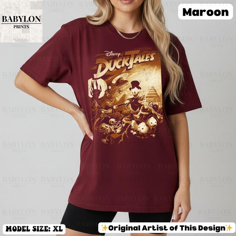 May include: Maroon t-shirt featuring a Disney DuckTales graphic. The shirt has a crew neck and short sleeves. The graphic is in shades of brown and tan. The text "Disney DuckTales" is at the top of the graphic. The model is wearing a size XL.