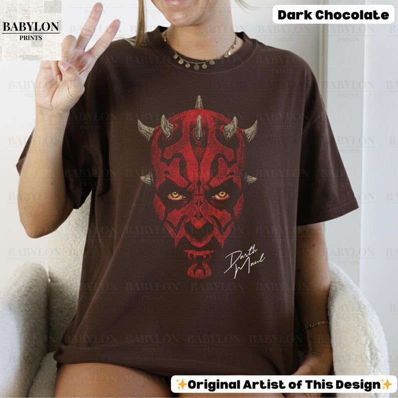Może przedstawiać: Ciemnobrązowy t-shirt w kolorze ciemnej czekolady z czerwoną grafiką Darth Maul. Grafika przedstawia szczeg&oacute;łowy projekt twarzy z rogami i napisem "Darth Maul". Koszulka jest z Babylon Prints.