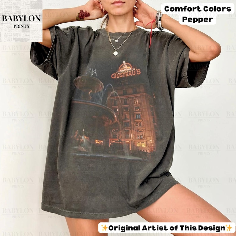 May include: A dark grey t-shirt featuring a graphic of a fountain and building, with the orange text "GUSTEAU'S". The shirt also has the text "Comfort Colors Pepper" and "Original Artist of This Design".