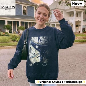 May include: A navy blue long-sleeve sweatshirt featuring a large, distressed graphic print of a white and blue clone trooper helmet on the front. The garment has a crew neckline, long sleeves with ribbed cuffs, and a ribbed hemline at the waist.