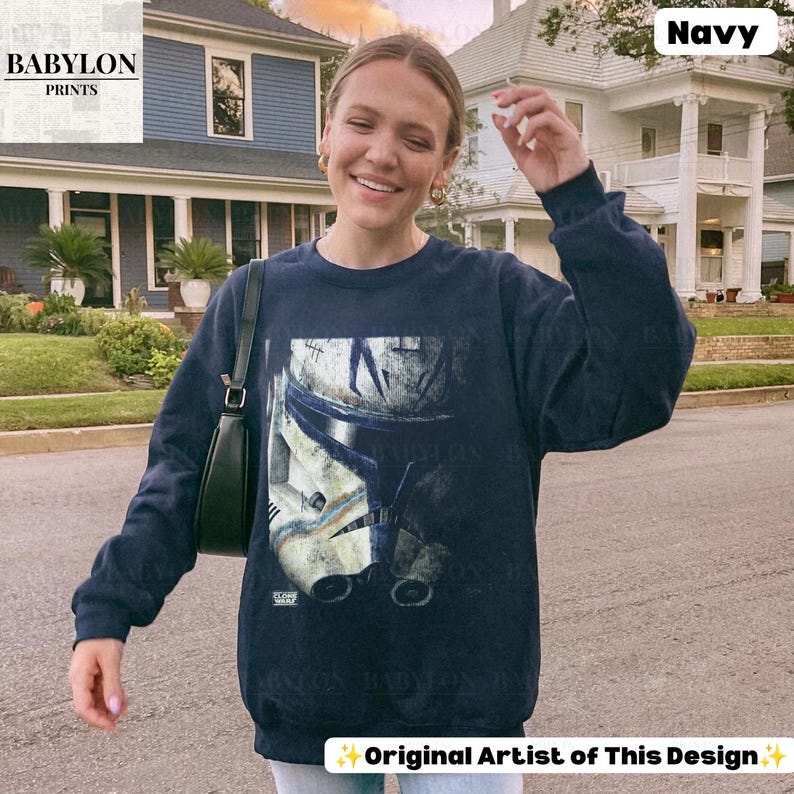 May include: A navy blue long-sleeve sweatshirt featuring a large, distressed graphic print of a white and blue clone trooper helmet on the front. The garment has a crew neckline, long sleeves with ribbed cuffs, and a ribbed hemline at the waist.