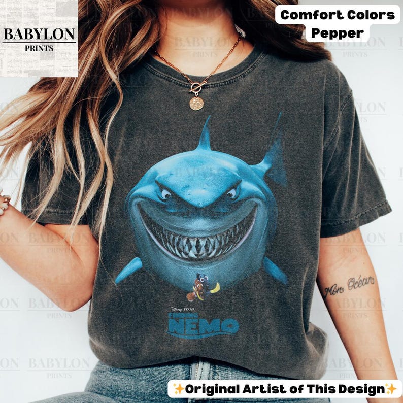 May include: A dark gray t-shirt featuring a large, blue cartoon shark with a wide grin. Below the shark is the text "Finding Nemo" with Nemo and Dory. The shirt has the text "Comfort Colors Pepper" at the top.