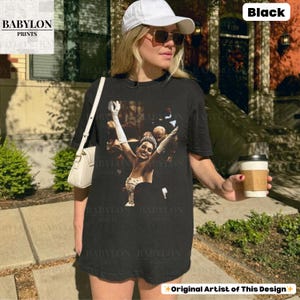 May include: Black t-shirt with a vintage image of a woman in a gown with her arms raised. The shirt has the text "BABYLON PRINTS" and "Original Artist of This Design". The model is wearing a white cap and sunglasses.