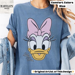 May include: Blue t-shirt featuring a Daisy Duck graphic. The cartoon character has a pink bow, eyelashes, and a yellow beak. The words "Daisy Duck" are written in cursive. The shirt is labeled "Comfort Colors" and "Original Artist of This Design."