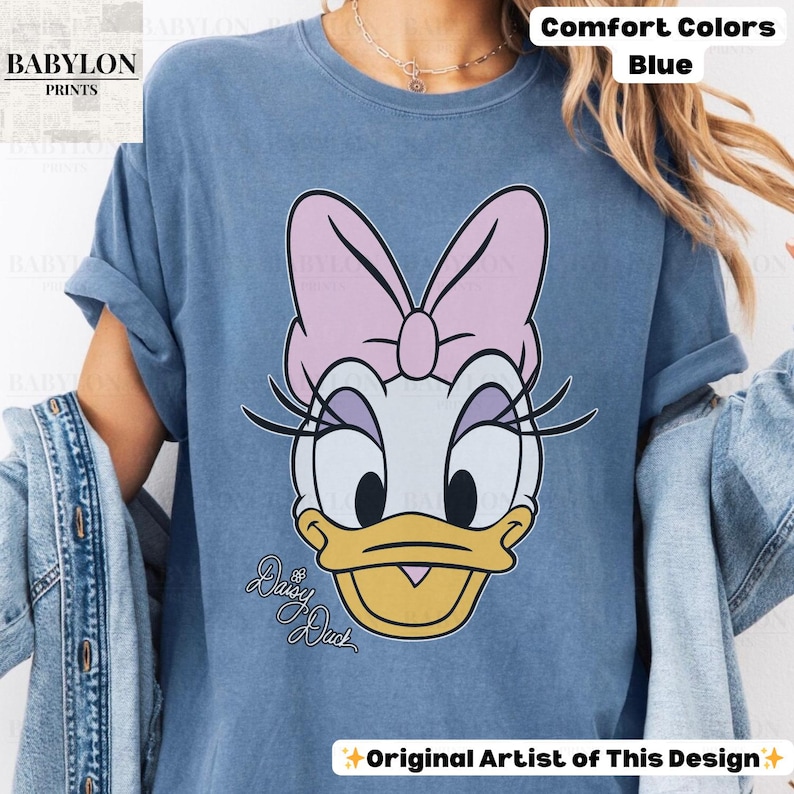 May include: Blue t-shirt featuring a Daisy Duck graphic. The cartoon character has a pink bow, eyelashes, and a yellow beak. The words "Daisy Duck" are written in cursive. The shirt is labeled "Comfort Colors" and "Original Artist of This Design."