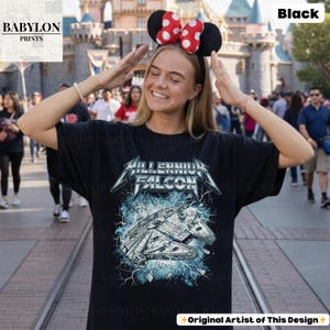 May include: Black t-shirt featuring a graphic of the Millennium Falcon spaceship with the text "MILLENNIUM FALCON" in a stylized font. The design includes blue lightning effects. The shirt is worn by a person wearing a Minnie Mouse ear headband.