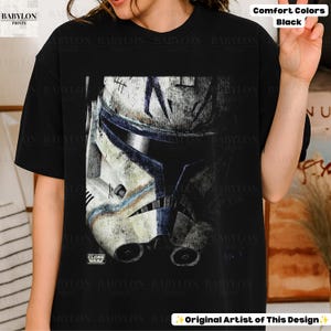 May include: A black t-shirt featuring a large, weathered graphic of a white and blue clone trooper helmet. The design includes the text "CLONE WARS" in the lower left corner of the graphic. The shirt has a crew neckline and short sleeves.
