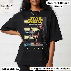 Puede incluir: Camiseta negra con un gr&aacute;fico de Star Wars: La Amenaza Fantasma. El dise&ntilde;o incluye el logotipo de Star Wars y una escena con dos personajes empu&ntilde;ando sables de luz. La camiseta es Comfort Colors y la modelo lleva la talla XL.