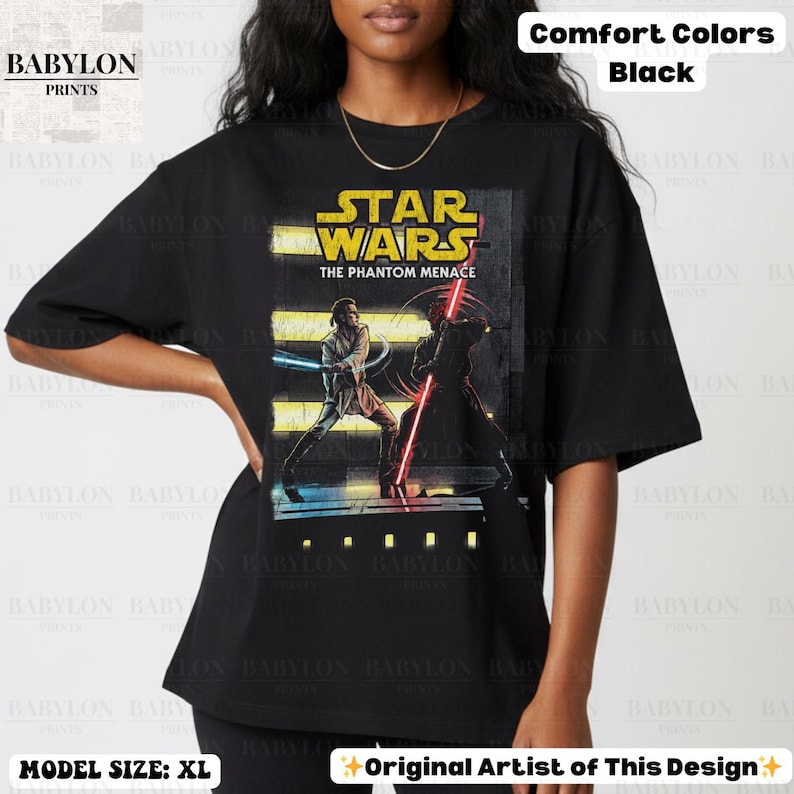 Puede incluir: Camiseta negra con un gr&aacute;fico de Star Wars: La Amenaza Fantasma. El dise&ntilde;o incluye el logotipo de Star Wars y una escena con dos personajes empu&ntilde;ando sables de luz. La camiseta es Comfort Colors y la modelo lleva la talla XL.