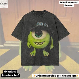 K&ouml;nnte beinhalten: Dunkelgraues, &uuml;bergro&szlig;es Premium-T-Shirt mit einer Cartoonfigur mit einem gro&szlig;en gr&uuml;nen Auge, die einen Hut mit den Buchstaben "MIT" tr&auml;gt. Das Shirt tr&auml;gt den Text "Premium Wash: Pepper" und "Oversized Premium Tee!"