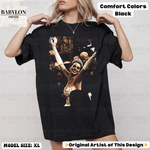 May include: Black Comfort Colors t-shirt with a graphic of a woman with her arms raised. The shirt has the text "BABYLON PRINTS" and "Original Artist of This Design". Model size is XL.