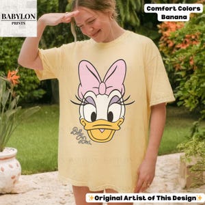 May include: A light yellow t-shirt featuring a Daisy Duck graphic with a pink bow and eyelashes. The shirt has the text "Baby Duck" and "Comfort Colors Banana". The shirt is a casual, oversized fit.