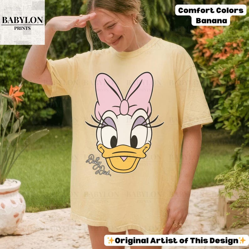 May include: A light yellow t-shirt featuring a Daisy Duck graphic with a pink bow and eyelashes. The shirt has the text "Baby Duck" and "Comfort Colors Banana". The shirt is a casual, oversized fit.
