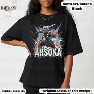 Może przedstawiać: Czarny t-shirt z grafiką Ahsoki Tano z mieczami świetlnymi i napisem "AHSOKA". Projekt zawiera pomarańczowe i niebieskie akcenty. Koszulka jest w stylu comfort colors.