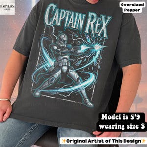 May include: Dark grey oversized t-shirt with a graphic of Captain Rex from Star Wars, firing a weapon with blue energy effects. The text "CAPTAIN REX" is in white and blue. The model is 175cm tall and wearing size S.