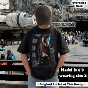 Może przedstawiać: Czarny, duży t-shirt z grafiką Obi-Wana Kenobiego z mieczem świetlnym. Koszulka zawiera napis "Obi-Wan Kenobi" i "Original Artist of This Design". Modelka ma 1,75 m wzrostu i nosi rozmiar S.