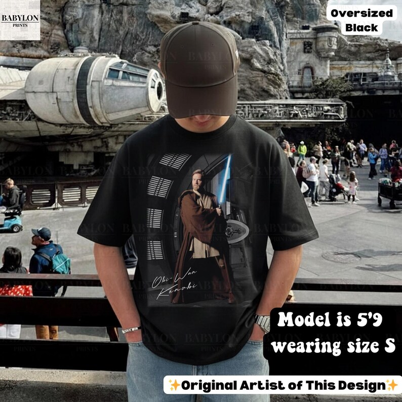 Może przedstawiać: Czarny, duży t-shirt z grafiką Obi-Wana Kenobiego z mieczem świetlnym. Koszulka zawiera napis "Obi-Wan Kenobi" i "Original Artist of This Design". Modelka ma 1,75 m wzrostu i nosi rozmiar S.