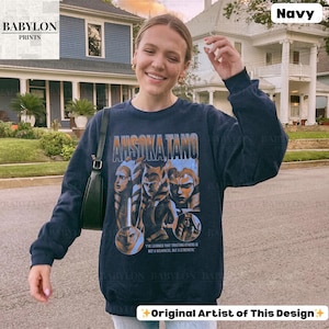 May include: Navy blue long-sleeved sweatshirt with a graphic print of characters from the Star Wars franchise. The text "Ahsoka Tano" is printed on the front of the shirt. The quote "I've learned that trusting others is not a weakness, but a strength" is printed below the graphic.