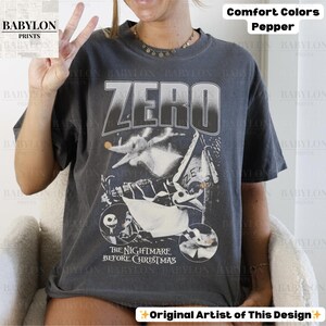 May include: Dark grey t-shirt featuring the word "ZERO" above a graphic from "The Nightmare Before Christmas". The design includes images of Zero, Jack Skellington, and other characters. The shirt is labeled "Comfort Colors Pepper".