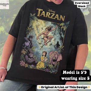May include: Black oversized t-shirt featuring a colorful graphic of Disney's Tarzan. The design includes Tarzan, gorillas, Jane, and an elephant. The shirt has the text "Disney Tarzan" in gold lettering. The text "Original Artist of This Design" is also visible.