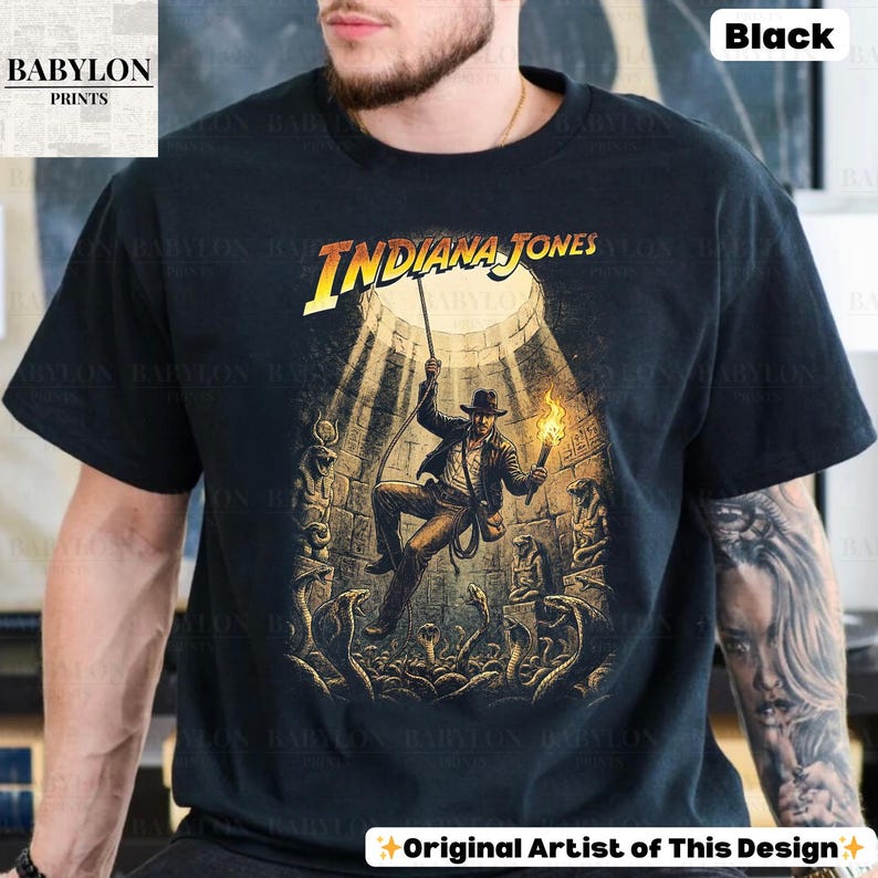 May include: Black t-shirt featuring a graphic of Indiana Jones in a temple setting, holding a torch and rope, with the text "Indiana Jones". The design includes snakes and stone carvings. The shirt also has the text "Original Artist of This Design".