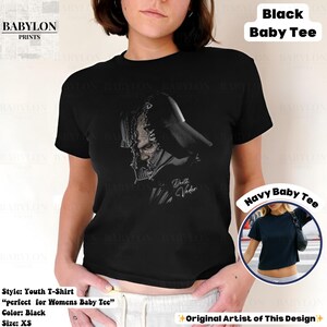 K&ouml;nnte beinhalten: Schwarzes Jugend-T-Shirt mit einer schwarz-wei&szlig;en Illustration von Darth Vader. Der Text "Darth Vader" ist kursiv geschrieben. Das Shirt ist im Baby-T-Shirt-Stil. Das Bild zeigt auch ein Navy Baby Tee.